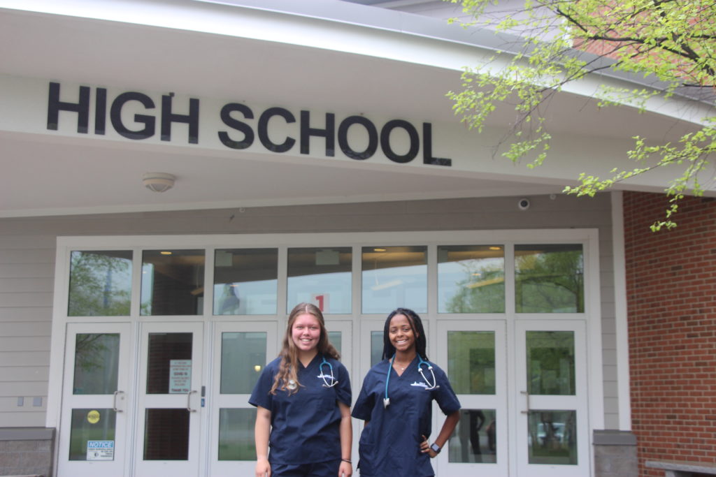 two female high school students in scrubs stand underneath high school sign